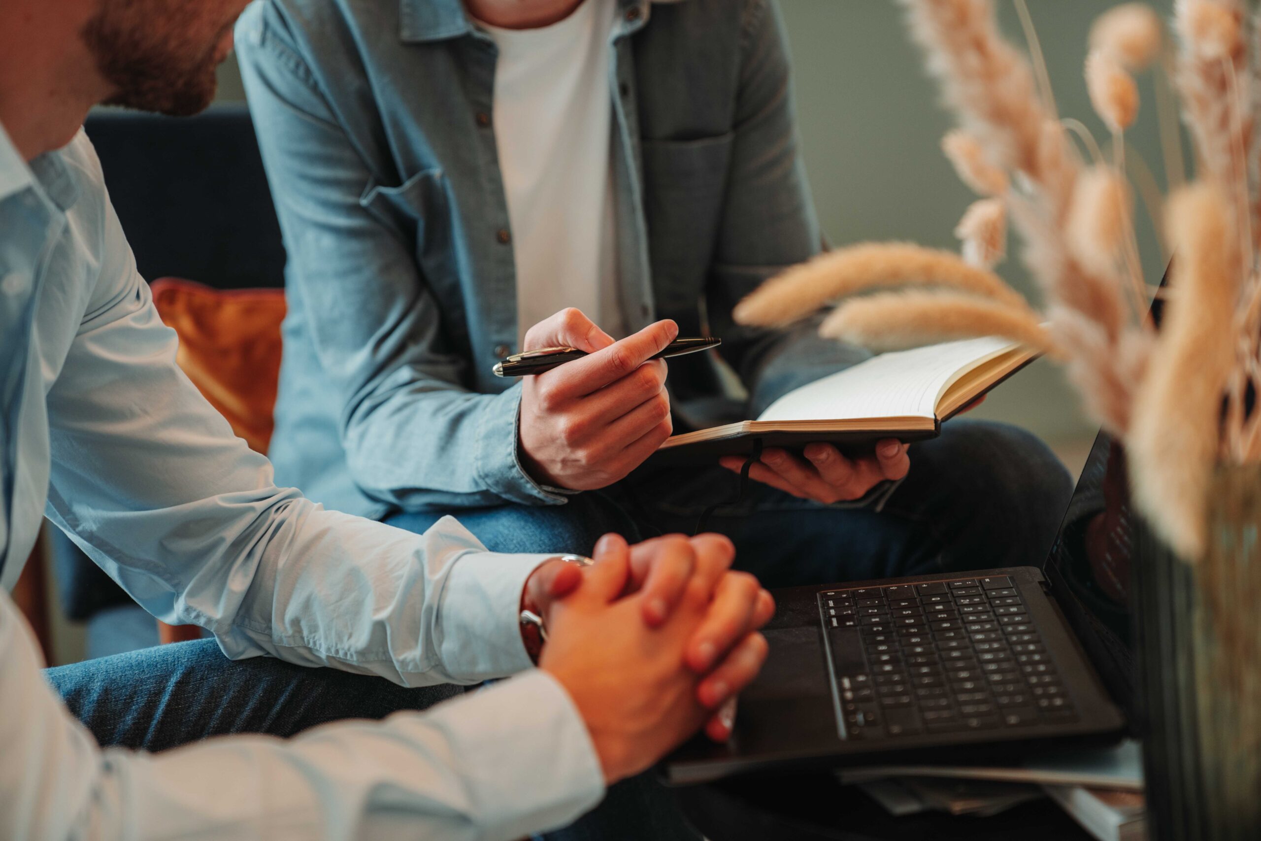 Close-up of two team members collaborating in a relaxed setting, with one holding a notebook and pen while discussing ideas. A laptop is open on the table, and dried pampas grass adds a decorative touch to the scene.